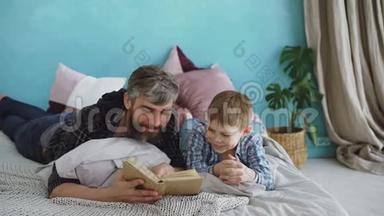 关爱的父亲正在给他的孩子读有趣的书，而男孩正在笑着和他的父母说话。 全尺寸床，明亮